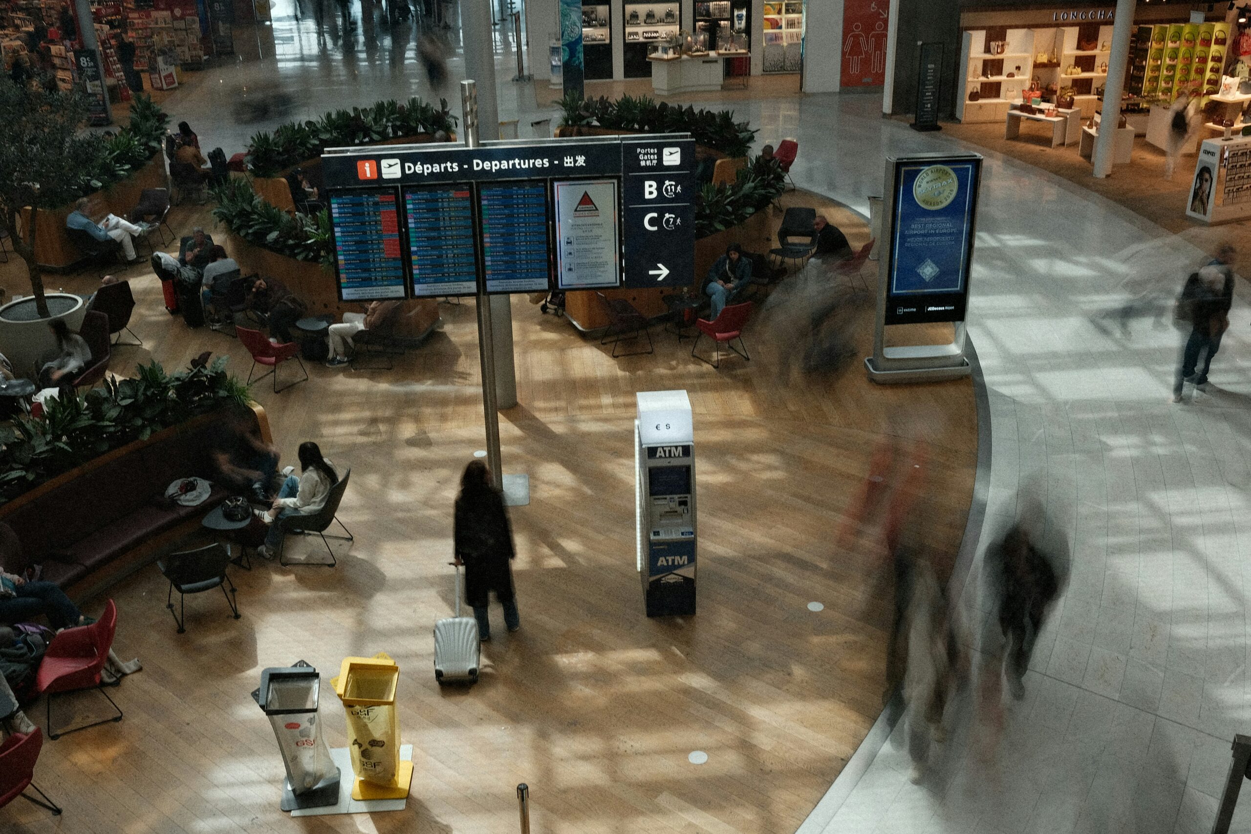 Airport departure area with travelers, luggage, flight information screens, and an ATM